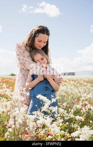 Eine Mutter umarmt sanft ihre Tochter zwischen blühenden Mohnblumen und Wildblumen auf einem sonnigen Frühlingsfeld und erschafft ein zartes Familienporträt unter einem Brig Stockfoto