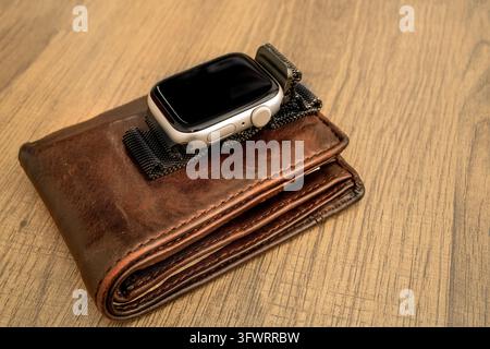 Eine Smartwatch mit dunklem Display und Stoffarmband liegt auf einem gefalteten Ledergeldbeutel auf einem Holztisch im Innenraum, ohne dass die Tageszeit angezeigt wird. Stockfoto