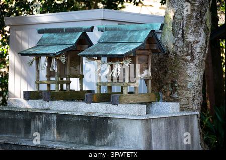 Zwei kleine hölzerne Shinto-Schreine (hokora) mit verwitterten grünen Metalldächern, Shimenawa-Seilen und Shide-Papierstreifen neben einem großen Baum in Japan. Stockfoto