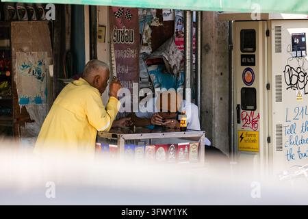 Chiang Mai, Thailand - 17. Januar 2026: Zwei Männer sitzen an einem kleinen Tisch und spielen Karten unter einem grünen Vordach auf einer Straße in Chiang Mai, Thailand. Stockfoto