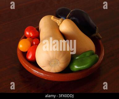 Organische Butternusskürbisse oder Butternuss-Squash, Auberginen oder Eierpflanzen, grüne Paprika und Tomaten in einer rustikalen Tonschale auf einem Holztisch. Stockfoto