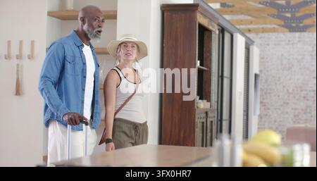 Vielfältiges Paar: Senior und erwachsene Frau stehen in der Küche, tragen Strohhut und halten einen Koffer Stockfoto