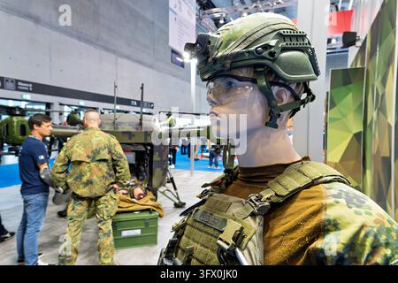 Berlin, 21.10.2026: Eine Schaufensterpuppe mit Gefechtshelm samt Halterung für ein Nachtsichtgerät, Schutzbrille, Plattenträger und Feldbluse im Flecktarnmuster steht am Stand der Bundeswehr auf dem Deutschen Kongress für Orthopädie und Unfallchirurgie DKOU 2026 im CityCube Berlin im Hintergrund stehen ein Soldat und ein Besucher vor der Rettungsdrohne Grille des Herstellers Avilus, in deren Ladeluke eine Übungspuppe liegt. Die unbemannte Drohne soll Verwundete künftig ohne Pilot aus dem Gefahrenbereich ausfliegen die Bundeswehr erprobt seit 2026 zwei Testmuster. Die zivil-militärische Zusamme Stockfoto