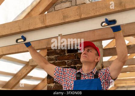 Bauarbeiter mit Handschuhen und roter Kappe misst einen Holzbalken präzise mit einer Wasserwaage, um während eines Bau- oder Renovierungsprojekts für Genauigkeit zu sorgen Stockfoto