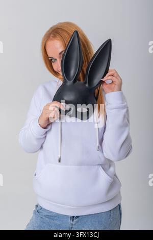 Junge Frau mit rotem Haar, die schüchtern hinter einer schwarzen Hasenmaske hervorlugt, einen weißen Hoodie und blaue Jeans trägt und eine Atmosphäre von Mystik und Verspieltheit erzeugt Stockfoto