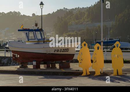 Traditionelles Fischerboot und gelbe Fischersilhouetten-Skulpturen zur Schau gestellt, Hauptstadt der Glasfische, L'Arena in San Juan de la Arena, Asturien, Spanien. Stockfoto
