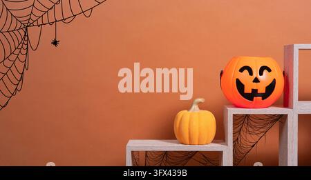 Vorderansicht eines orangefarbenen, kürbisförmigen Plastiktopfs und eines Keramik-Kürbisses auf einem Holzregal vor einem braunen Hintergrund mit Spinnweben. Stockfoto