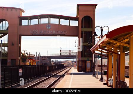 Eine Fußgängerbrücke, die als San Bernardino gekennzeichnet ist, überspannt Bahngleise neben einem leeren Bahnsteig mit orangefarbenen Unterständen, dekorativen Laternen und roten Signalen. Stockfoto