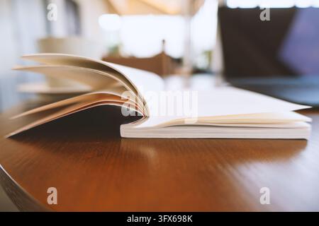 Ein aufgeschlagenes Hardcover-Buch liegt auf einem polierten Holzschreibtisch in einem sanft beleuchteten modernen Arbeitszimmer, mit einem Laptop und einer Kaffeetasse im Hintergrund unscharf, was eine ruhige Atmosphäre konzentrierten Lernens und intellektueller Arbeit vermittelt Stockfoto