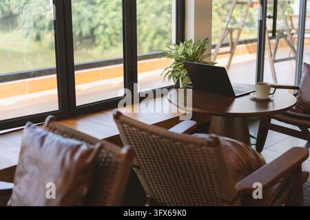 Gemütliches modernes Café-Interieur mit einem schwarzen Laptop, der auf einem runden Holztisch geöffnet liegt, neben einer Keramik-Kaffeetasse mit Untertasse, flankiert von zwei gewebten Korbstühlen mit Lederpolstern; große bodengleiche Fenster zeigen üppige Grünpflanzen und ein ruhiges Wasserweg draußen, was auf eine ruhige städtische oder vorstädtische Umgebung in Südostasien, möglicherweise Chiang Mai oder Hoi An, hindeutet, wobei natürliches Tageslicht die warmtonige minimalistische Dekoration erhellt. Stockfoto
