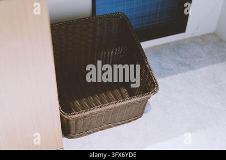 Leerer rechteckiger Weidenaufbewahrungskorb, der auf hellem grauem Fliesenboden neben einem Holzschrank und einem Fenster mit blauem Gittermuster platziert ist, moderne minimalistische Inneneinrichtung Stockfoto