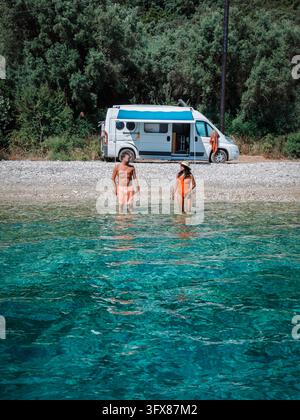 Zwei Reisende waten am klaren Ufer neben einem geparkten Wohnmobil auf Lefkada, Griechenland, und genießen einen hellen Sommertag inmitten türkisfarbenen Wassers und grünem C Stockfoto