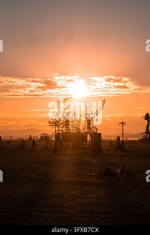 Eine große Drahtskulptur, die Figuren darstellt, steht während des Sonnenuntergangs auf einer Wüstenebene, während entfernte Radfahrer über die flache, trockene Landschaft fahren unter einem Stockfoto