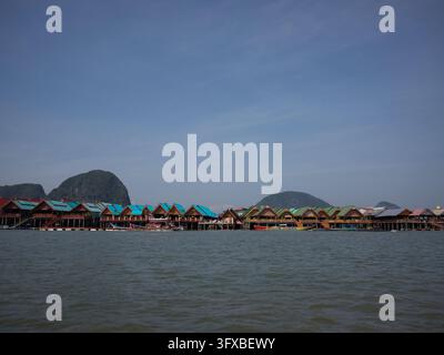 Reihe farbenfroher Holzstiltshäuser mit blaugrünen und grünen Dächern entlang des Ufers unter Kalksteinhügeln und einem blauen Himmel in der Phang-Nga-Bucht, Thailand. Stockfoto