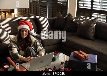 Erwachsene Frau mit Weihnachtsmannmütze sitzt auf dem Boden im Wohnzimmer und packt Geschenke in der Nähe eines Laptops. Gemütlich, Feiertag, Verpackung, Geschenk, Computer, Sofa, gestreift Stockfoto