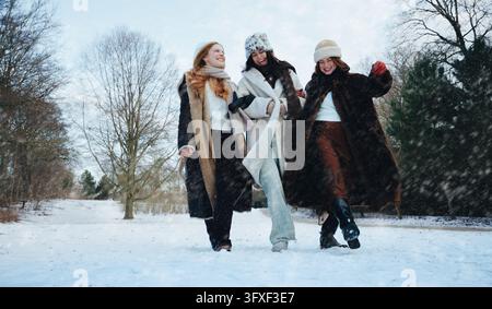 Drei junge Frauen gehen lachend zusammen durch den Schnee in einem Winterpark und feiern Freundschaft, Outdoor-Lebensstil und einen fröhlichen Tag mit Freunden Stockfoto