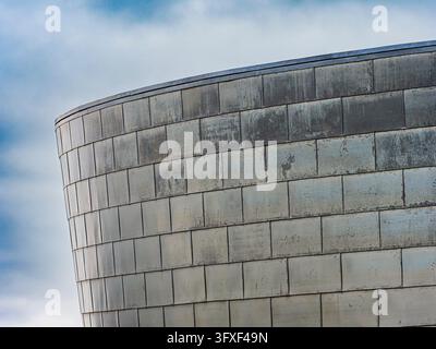 Modernes Architekturgebäude mit einer gebogenen Metallfassade, ausgestattet mit verwitterten rechteckigen Fliesen, vor einem bewölkten Himmel Stockfoto