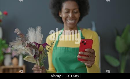 Floristin mit grüner Schürze hält Smartphone und Strauß aus getrockneten Gräsern und Eukalyptus im Blumengeschäft-Studio; Freude an kleinen Unternehmen. Stockfoto
