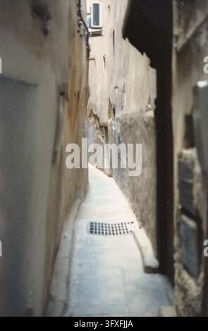 Schmale Stein-Gasse zwischen verwitterten alten Gebäuden, ein Metall-Abflussgitter im gepflasterten Boden und verschwommene Wände, die den Blick einrahmen. Stockfoto