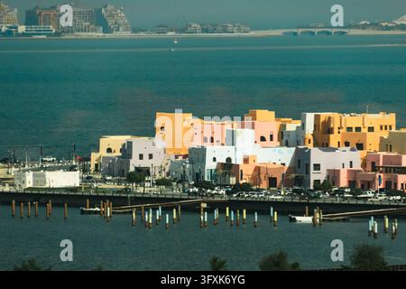 Helle Ansicht der pastellfarbenen Gebäude im Mina-Distrikt am Alten Hafen von Doha mit blauem Meerwasser und entfernter Küstenarchitektur an einem sonnigen Tag. Stockfoto