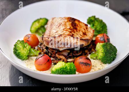 Gegrilltes Fischfilet serviert über sautierten Pilzen mit Brokkolischoten, gerösteten Kirschtomaten und cremiger Sauce in einer weißen Schüssel Stockfoto
