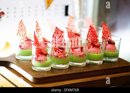 Gourmet-Erdbeermousse-Dessert in kleinen Glasbechern mit grüner Pandanschicht und roter Zuckerdekoration auf Holzservierplatte. Verschiedene Mini-Parfaits Stockfoto