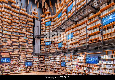 Tokio, Japan, 22. NOV 2024 – Ein weitläufiger Innenraum zeigt bodentiefe Holzregale, die mit Büchern gefüllt sind, abwechselnd mit blauem Digitalbildschirm Stockfoto