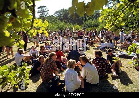 Junge Erwachsene entspannen sich, unterhalten sich und trinken zusammen auf einer grasbewachsenen Parkwiese während eines sonnigen Outdoor-Festivals. Das Treffen zeigt Geselligkeit, Freizeit Stockfoto