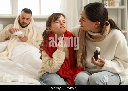 Familien-Gesundheitsversorgung und Grippesaison, Mutter kümmert sich um ihre kranke Tochter mit Halsschmerzen, während der Vater mit Erkältungssymptomen im Bett ruht, zu Hause im Schlafzimmer Stockfoto