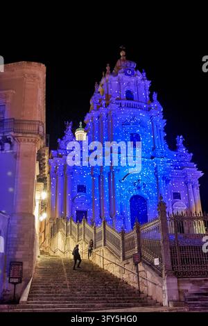 Blaues Licht projiziert auf die barocke Fassade der Kirche San Giorgio in Ragusa Ibla, Italien, während zwei Personen die steinernen Treppen daneben hinaufsteigen. Stockfoto