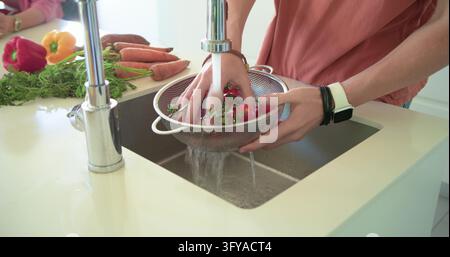 Haushaltsmitglieder spülen Radieschen in einem Sieb unter dem Wasserhahn am Küchenwaschbecken, Smartwatch sichtbar. Produkte, Gemüse, Waschen, Karotten, Paprika, Duo, Cou Stockfoto