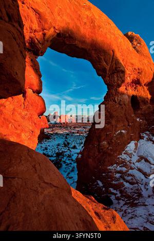 Ein Winterblick auf die schneebedeckten La-Sal-Berge, eingerahmt durch die natürliche Sandsteinöffnung des Turret Arch im Arches National Park, Utah, USA. Die markante geologische Formation zeigt rote Felsnasen und Bögen, die durch die Erosion des Colorado-Plateaus gekennzeichnet sind. Schnee bedeckt das Wüstengelände und die umliegenden Felsstrukturen, im Kontrast zum satten Terrakotta-Sandstein unter einem klaren Himmel. Dieses dramatische natürliche Fenster zeigt die landschaftliche Schönheit und Geologie des amerikanischen Südwestens. Eine optimierte Version eines Bildes des US National Park Service. Credit: NPS Stockfoto