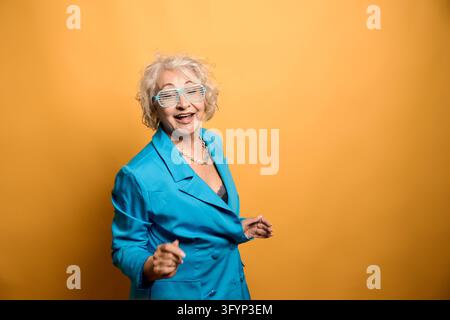 Fröhliche Seniorin hat Spaß beim Tanzen, trägt Sonnenbrille und einen blauen Blazer, drückt Freude auf einem gelben Hintergrund aus Stockfoto
