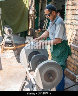 Osh, Kirgisistan – 19. Mai 2022: Handwerker mit einer Kappe und grüner Schürze schärft eine Klinge an einem Schleifrad, wobei Funken in seiner Werkstatt fliegen Stockfoto