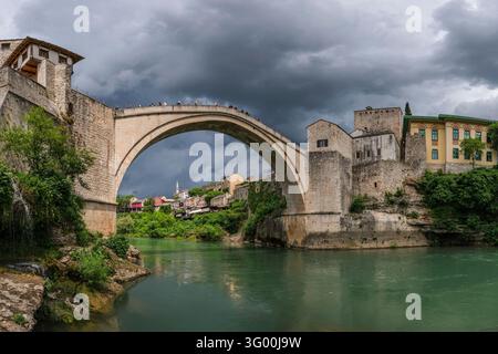 Dunkle Gewitterwolken sammeln sich über der Alten Brücke in Mostar, während Besucher über ihr erhöhtes steinernes Kopfsteinpflaster gehen. Stockfoto