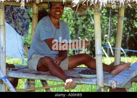 männliche Arbeiter lächelnd sitzt in seinen Schutz ein Vogel erschrecken Gerät in einem Reisfeld in Kerobokan Bali Indonesien Stockfoto