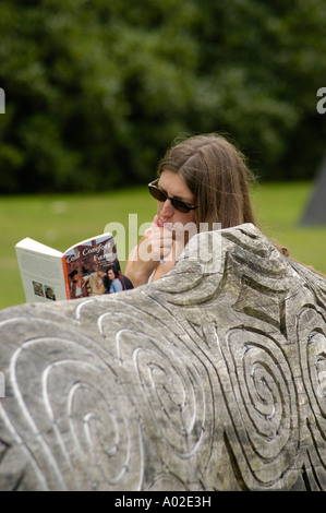 Junge Frau trägt Sunglassses Cold Comfort Farm Taschenbuch lesen sitzen auf geschnitzte Holzbank Machynlleth, Powys wales Stockfoto