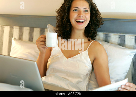 Frau Mit Laptop Und Zeitung Im Bett, Frau mit Laptop und eine Tasse Kaffee im Bett Stockfoto
