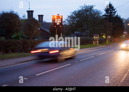 eine blinkende Geschwindigkeit Warnzeichen und schnell bewegenden Feierabendverkehr durch das Dorf große Barton in Suffolk, UK Stockfoto