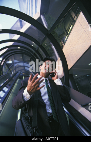 Asiatische amerikanische Geschäftsmann über sein Handy und gestikulierend, wie er reitet und outdoor Rolltreppe nach unten Stockfoto