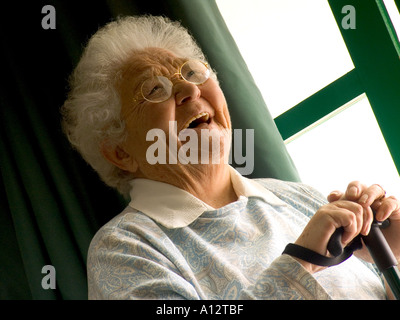 Ältere, fröhliche, unabhängige, ältere, ältere Dame, die mit einem Spazierstock am Fenster in ihrem hellen, modernen Zuhause sitzt Stockfoto
