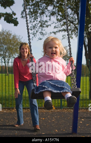 Vertikale Porträt einer jungen kaukasischen Mutter schob ihre blonde Mädchen auf einer Schaukel im Park. Stockfoto