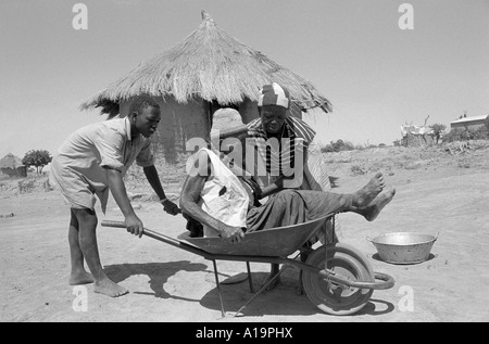 B/W eines Jungen, der seine ältere Großmutter auf den Dorfmarkt in einer Schubkarre in der Nähe von Zaka, Simbabwe, nimmt. Stockfoto