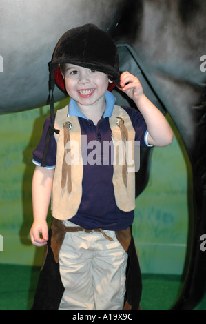 Cute toothy Lächeln eines Jungen in Reiten Kleidung vor ein Pferd Statue bei einem Kindermuseum gekleidet Stockfoto