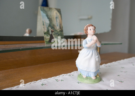 Statuette der ein junges Mädchen im weißen Kleid auf einem bestickten Tischdecke mit einem Gemälde der Mutter Mary hinter Stockfoto