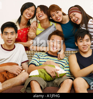 Nahaufnahme von drei jungen Männern, die mit vier Frauen, die hinter ihnen stehen auf einer Couch sitzend Stockfoto