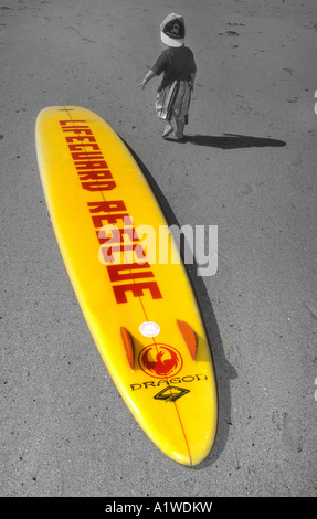 Lifeguard Rescue Surfbrett liegend auf einem Sandstrand In Newquay Cornwall, mit einem kleinen Jungen zu Fuß vorbei. Stockfoto