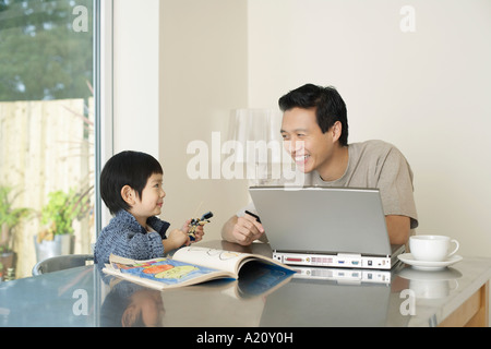 Vater und Sohn sitzen am Küchentisch zusammen; Laptop und Malbuch auf Tisch Stockfoto