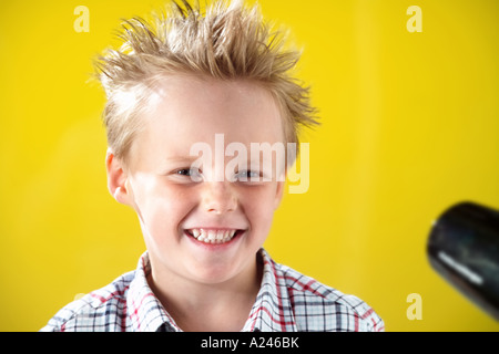 Blonde junge im Alter von 5-7 Jahren gegen gelbe Studio-Hintergrund lustige Grimassen und dabei seine Haare Schlag getrocknet. Stockfoto