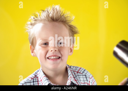 Blonde junge im Alter von 5-7 Jahren gegen gelbe Studio-Hintergrund lustige Grimassen und dabei seine Haare Schlag getrocknet. Stockfoto